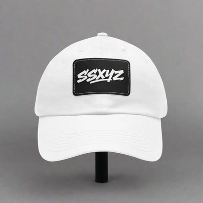 White dad hat with a black rectangular patch on the front displaying the white text "SSXYZ", the shorthand logo for Stylesavvy.xyz. The hat has a curved brim and an adjustable back strap.